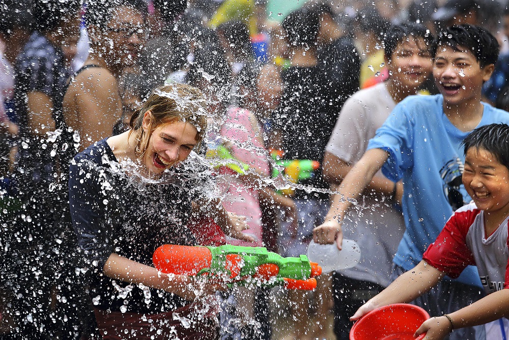 A joyful Songkran celebration in Pattaya, Thailand, featuring people splashing water, laughing, and playing with water guns. A woman with a water gun is being drenched while children and locals enjoy the festival, highlighting the excitement of the Wan Lai Festival on April 19.